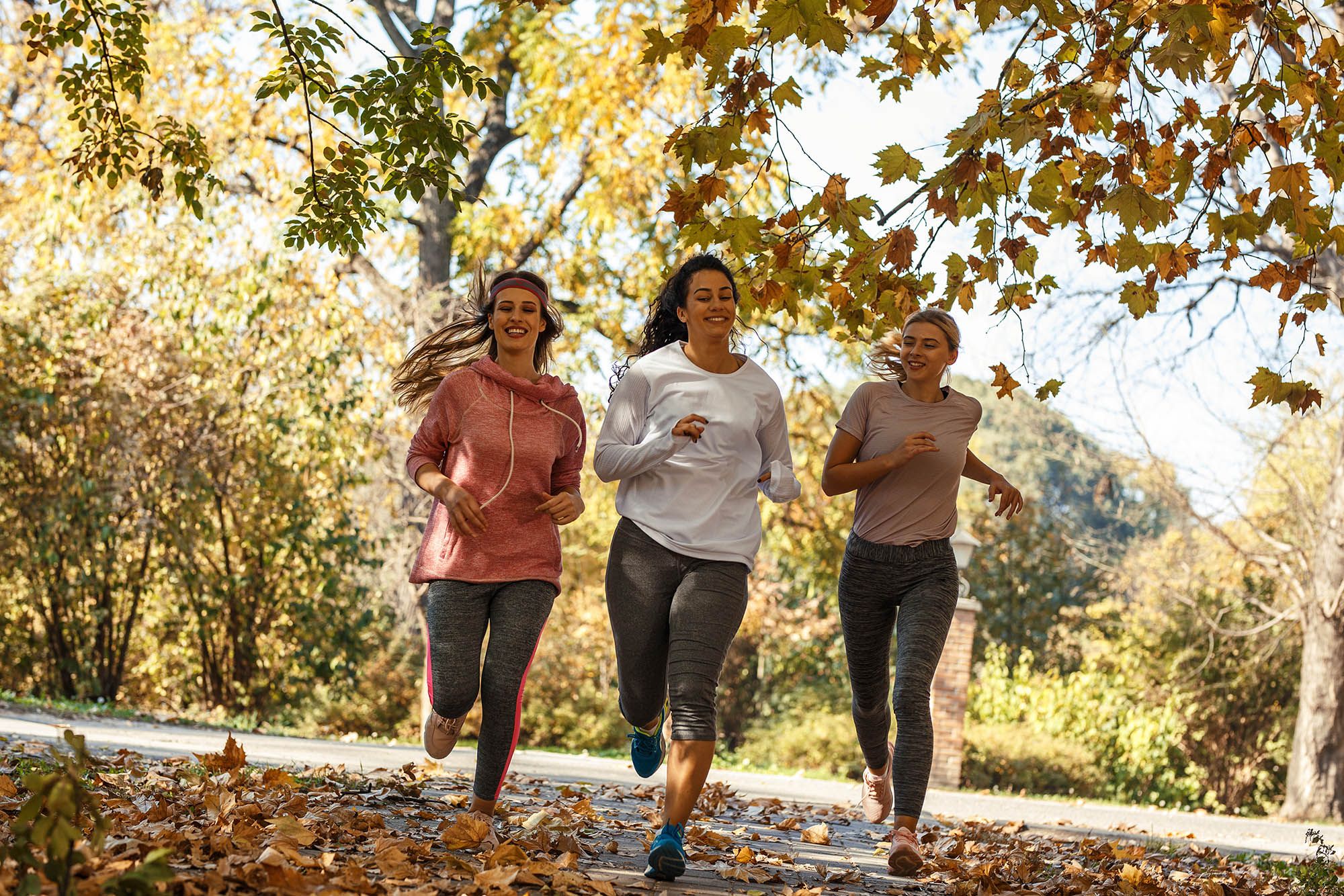 Three women running in workout gear through autumn park path